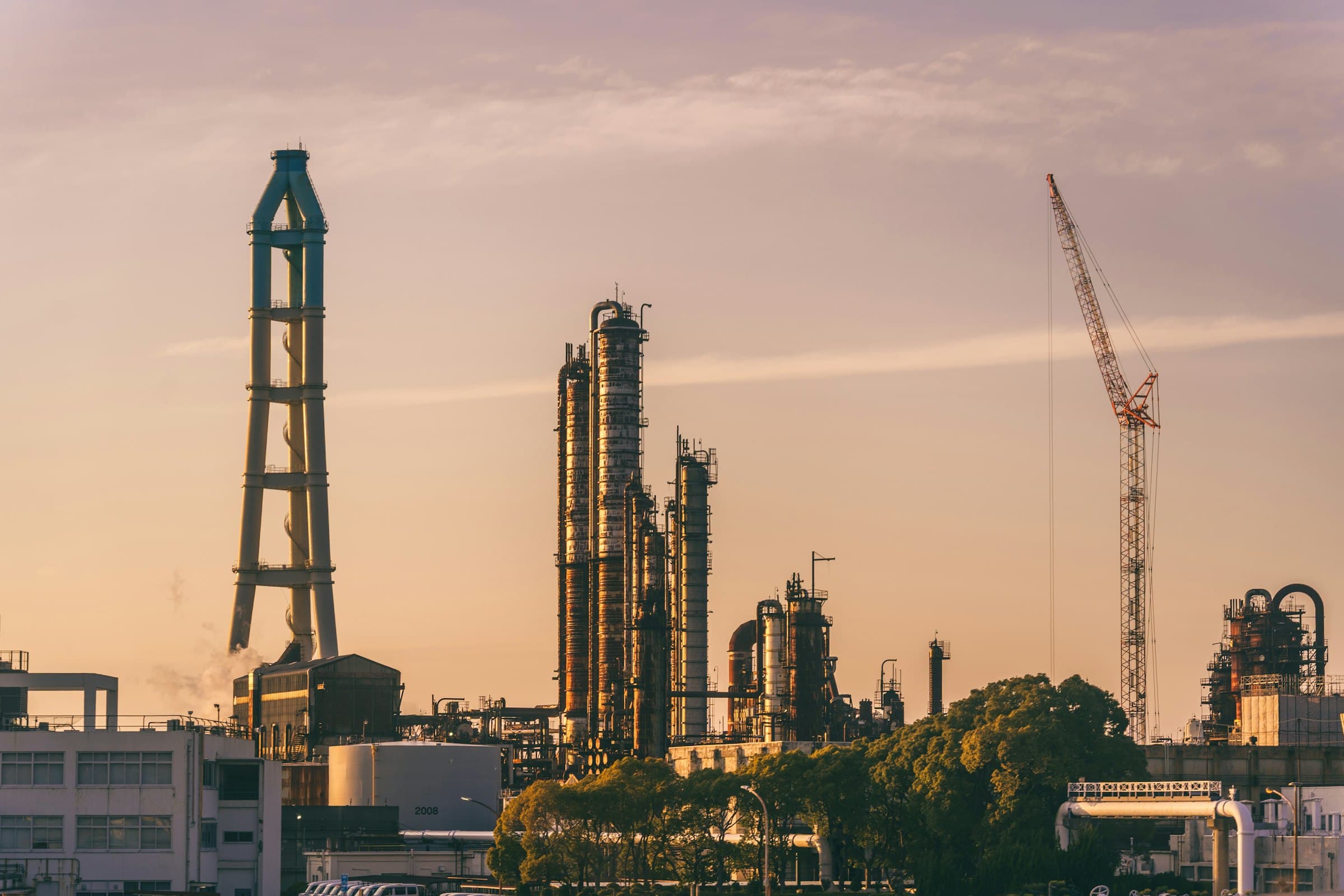 Refinery towers and a construction crane against a warm sky at dusk